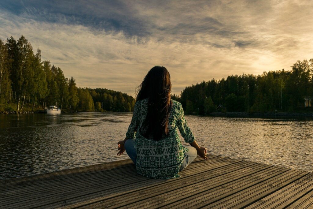Entspannungsmethoden: Eine Frau im Meditations-Sitz auf einem Holzsteg mit Blick auf einen See. 