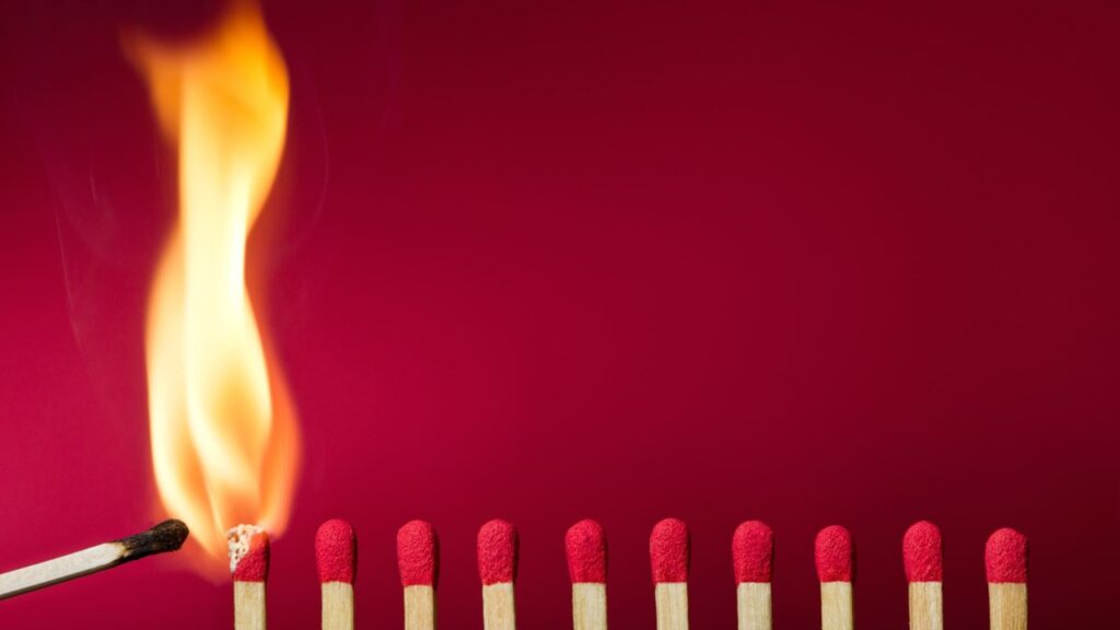 Eine Reihe frischer Streichhölzer mit rotem Kopf nebeneinander. Das erste Streichholz wird mit einem brennenden Streichholz angezündet und eine helle Flamme züngelt auf. Zweck des Bildes ist es, das Burnout-Syndrom am Beispiel brennender Streichhölzer darzustellen.