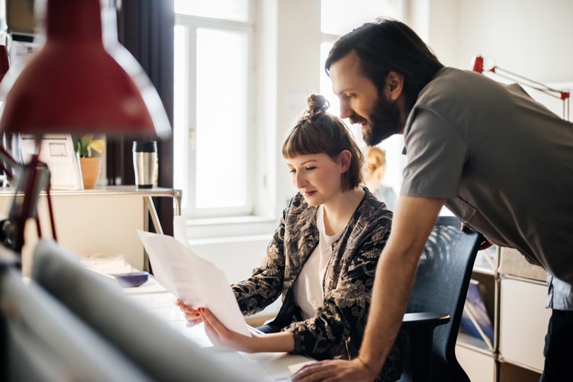 Achtsamkeit am Arbeitsplatz: Eine Frau und ein Mann in Arbeitsumgebung im Büro am Schreibtisch, die sich gemeinsam ein Schriftstück anschauen.