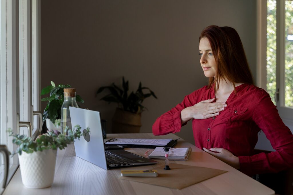 Achtsamkeit im Home-Office: Frau sitzt am Schreibtisch vor aufgeklapptem Notebook. Ihre rechte Hand liegt flach auf ihren Brustkorb, die linke Hand seitlich am Rippenbogen, um ihre Atmung zu spüren.