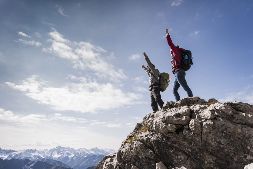 Ein Mann und eine Frau in Wanderkleidung und mit Rucksack stehen auf einem Berggipfel uns reißen freudig ihre Arme in die Höhe.